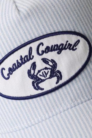 Close-up of a cap with 'Coastal Cowgirl' and crab logo on a light blue striped background