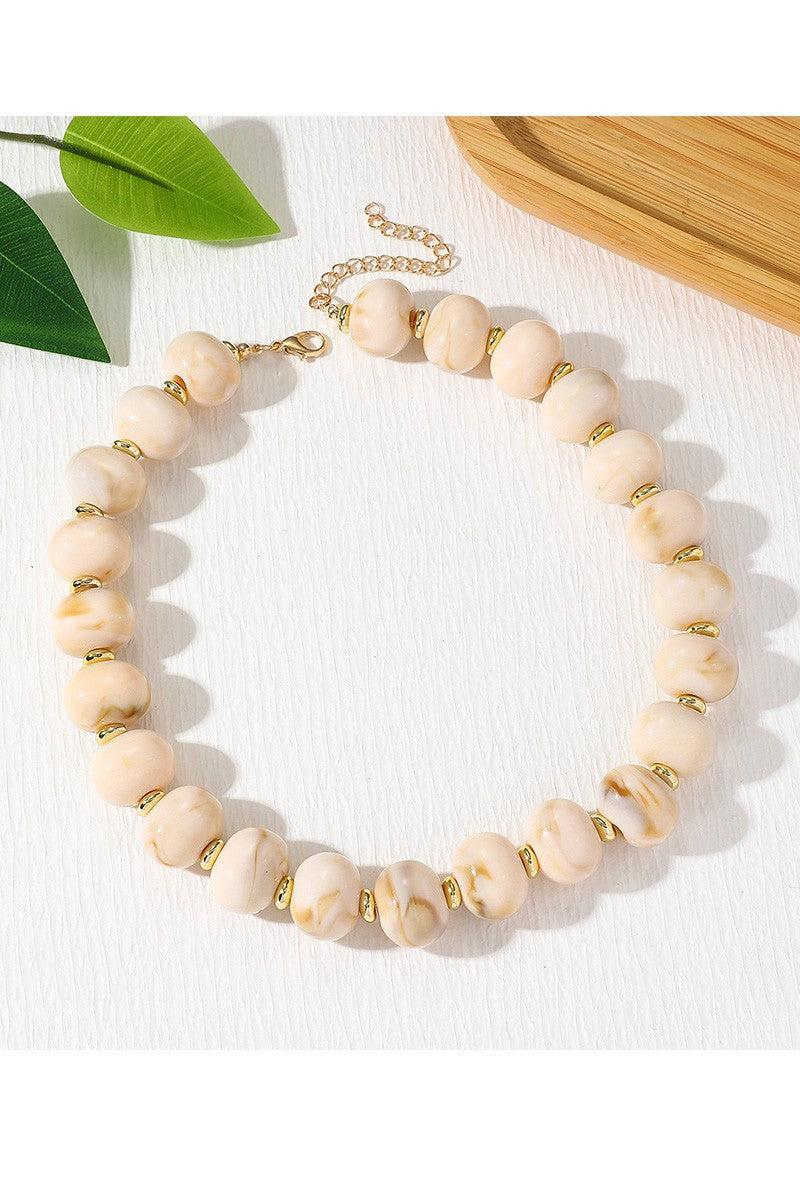 A close-up of a beaded strand with large round pale blush-beige stones separated by small metallic gold spacers, photographed on a white surface.