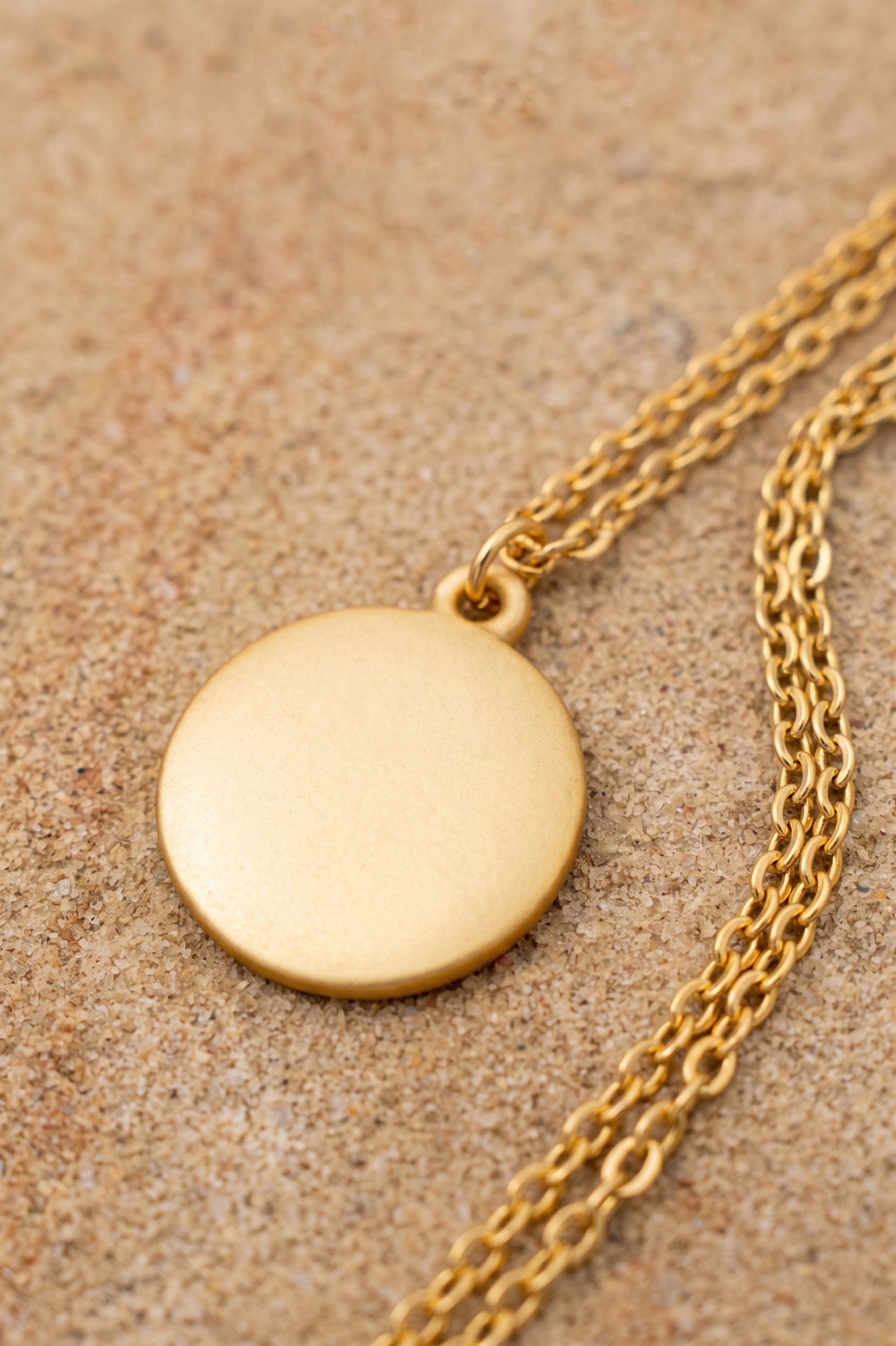 A gold disc pendant necklace with a matching chain, displayed on a textured beige sand-like surface.