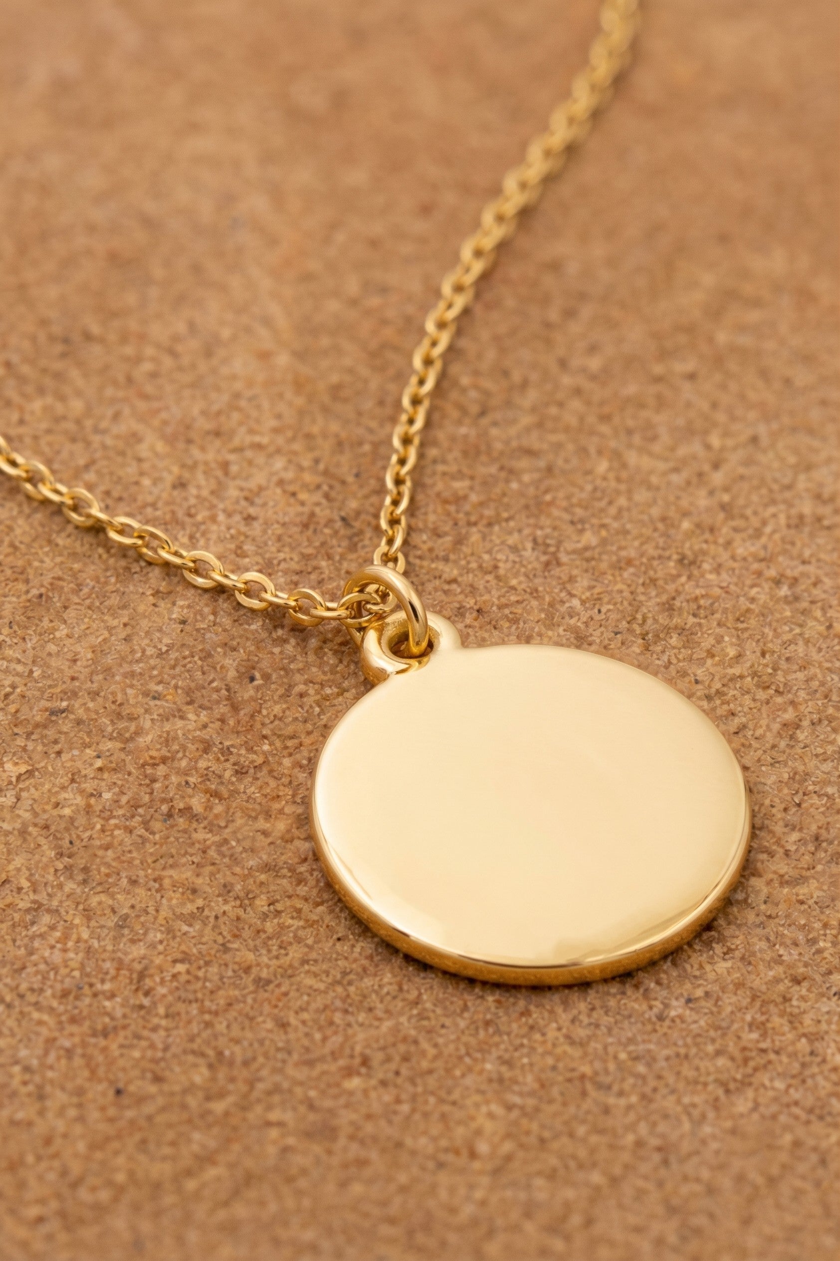 A gold disc pendant necklace on a matching gold chain, displayed on a textured brown background.