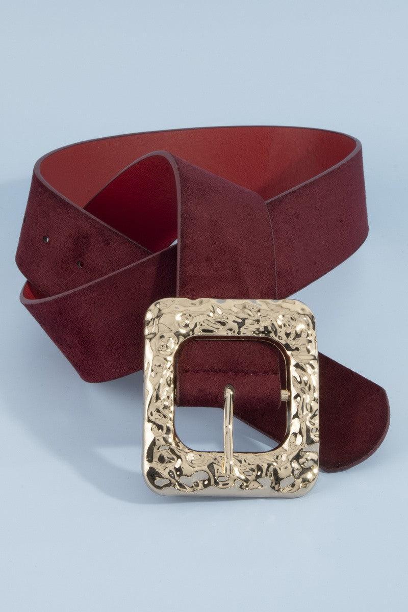 A rippled square frame buckle with a molten, hammered gold-tone finish sits front and center, on a burgundy faux suede belt.