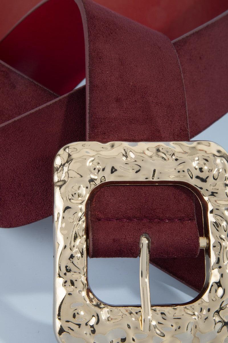 A rippled square frame buckle with a molten, hammered gold-tone finish sits front and center, on a burgundy faux suede belt.
