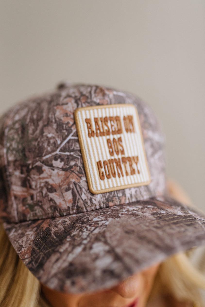 A close-up of a person wearing a camouflage baseball cap with a rectangular front patch that reads 'Raised on 90s Country' in brown text on a cream striped background.