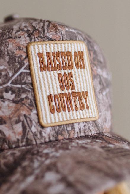 Close-up of a camouflage baseball cap with a rectangular embroidered patch reading 'RAISED ON 90S COUNTRY' in brown lettering on a cream striped background.