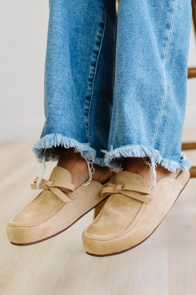 A person sitting on a wooden stool wearing beige suede slip-on loafers with bow details and frayed blue jeans.