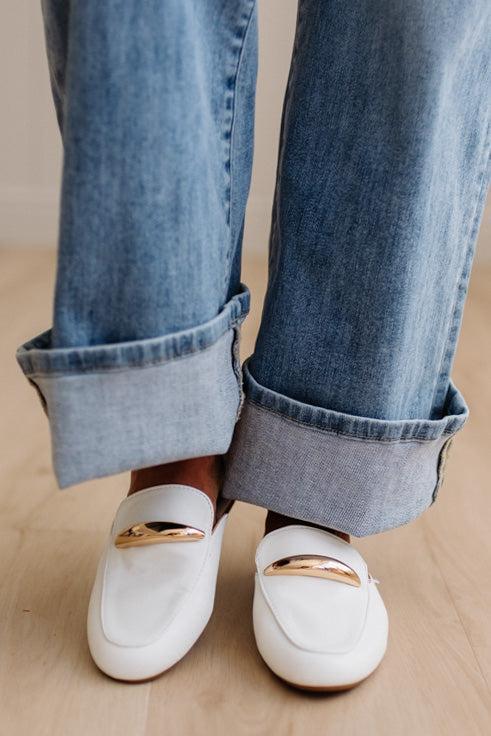 White loafers with gold accents worn with rolled-up blue jeans on a wooden floor.