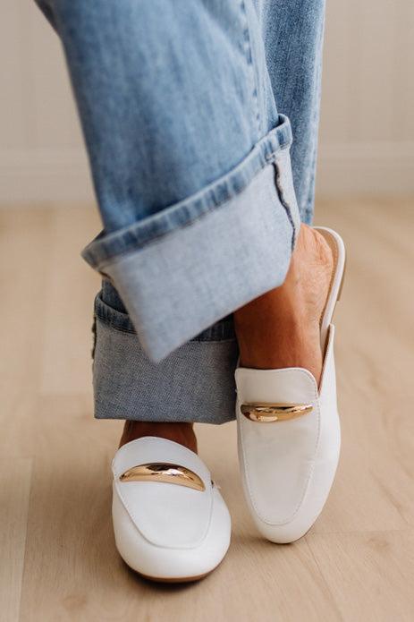 White loafers with gold accents worn with rolled-up jeans on a light wooden floor.