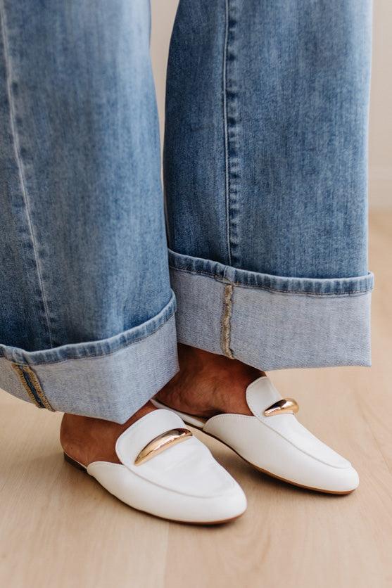 White loafers with gold accents worn with rolled-up blue jeans on a neutral background