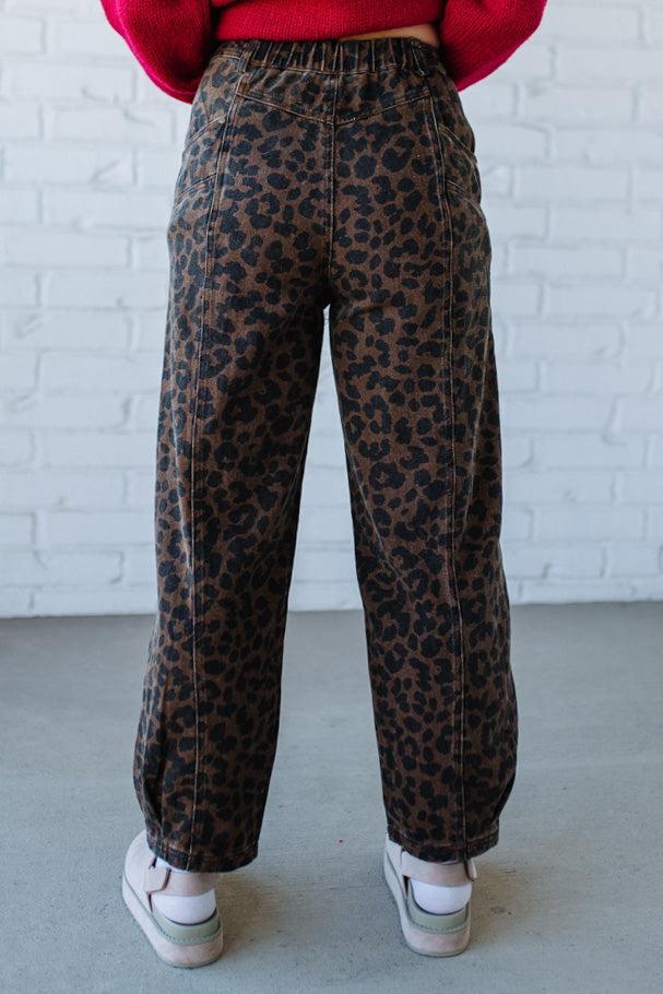 Person wearing leopard print pants with a red sweater and white sneakers against a white brick wall.