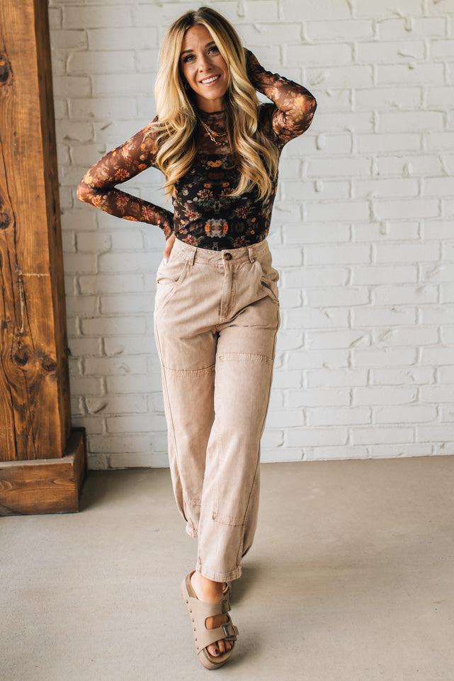 Woman wearing a floral top and beige pants against a white brick wall.