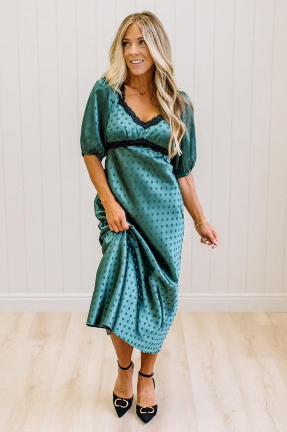 A woman modeling a teal green midi-length slip dress with a black polka dot pattern, black lace trim at the V-neck, and puff sleeves, paired with black heels, standing in a room with white paneled walls and light wood floors.