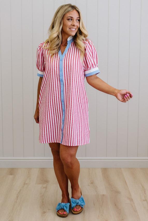 Woman wearing a red and white striped dress with blue accents against a white paneled wall.