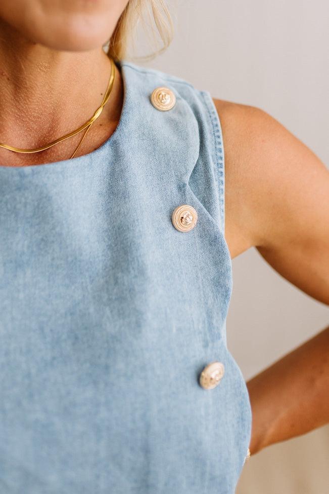 Close-up of a person wearing a light blue sleeveless top with decorative beige buttons along the shoulder strap, paired with a thin gold necklace against a soft neutral background.
