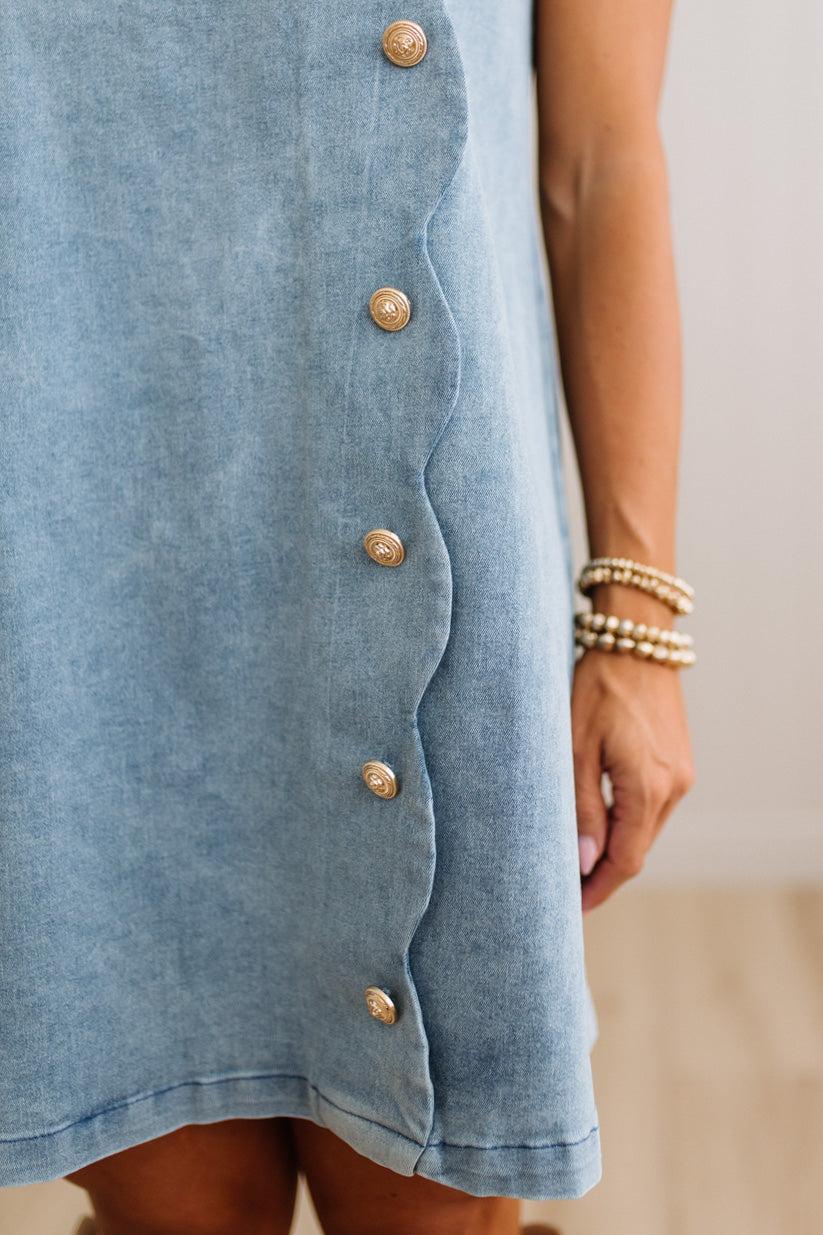 Close-up of a light blue denim dress with a scalloped edge and gold-tone buttons, worn by a model whose arm and hand are visible against a soft beige background.