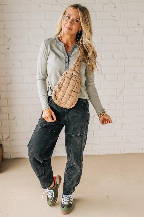 Woman wearing a gray long-sleeve shirt, dark jeans, and green sneakers, holding a beige crossbody bag against a white brick wall.