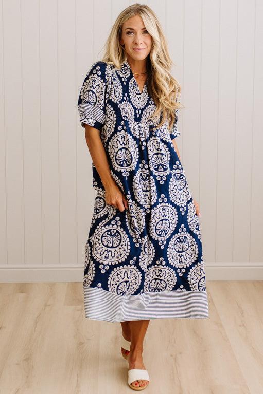 A woman modeling a navy blue and white patterned midi dress with short sleeves, a V-neckline, and a striped hem, standing in front of a white paneled wall and light wood floor.