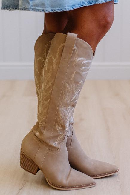 Beige cowboy boots worn with a denim skirt on a light wooden floor.