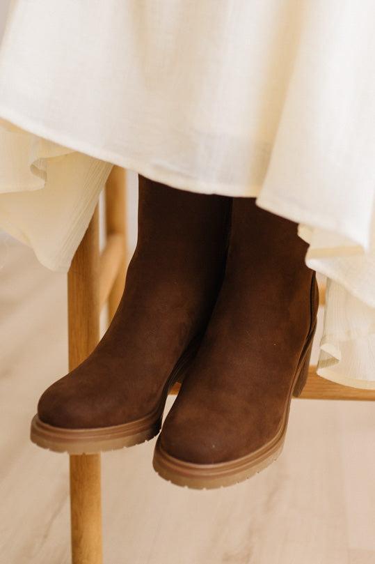 A pair of dark brown suede ankle boots with tan soles, shown on a wooden chair beneath a cream-colored dress.