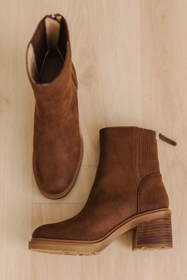 A pair of brown suede ankle boots with elastic side panels and stacked block heels, displayed on a light wood floor.