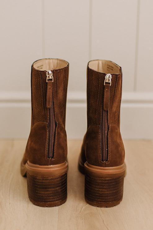 A pair of brown suede ankle boots with side zippers and stacked block heels, shown from the back on a light wood floor against a white paneled wall.