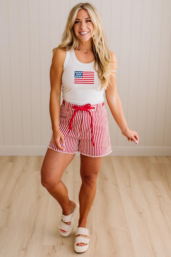 Woman wearing a white tank top with an American flag design and red and white striped shorts on a wooden floor.