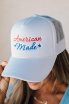 Blonde woman wearing a white mesh back trucker hat with script letters in red and blue emboidered "American Made" at the front.
