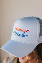 Blonde woman wearing a white mesh back trucker hat with script letters in red and blue emboidered "American Made" at the front.
