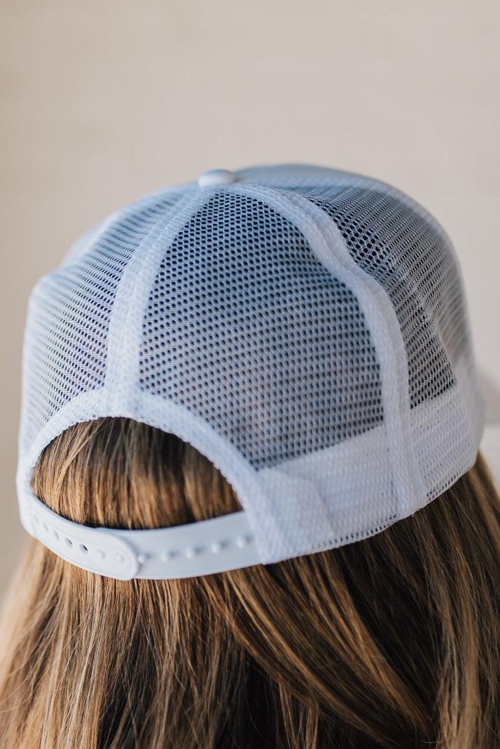 Blonde woman wearing a white mesh back trucker hat with script letters in red and blue emboidered "American Made" at the front.