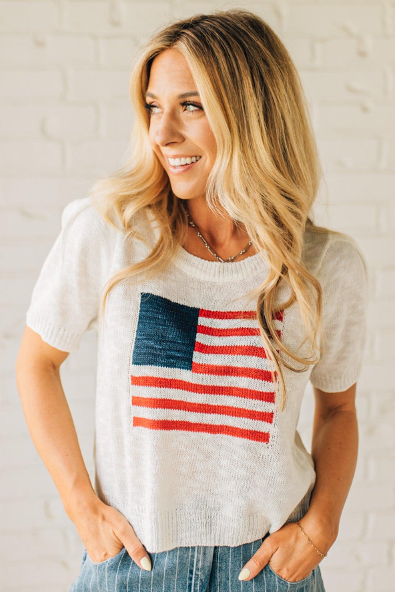 BLONDE WOMAN WEARING A WHITE AMERICAN FLAG TOP WITH SHIRRING DETAILS SHORT SLEEVES SWEATER TOP.