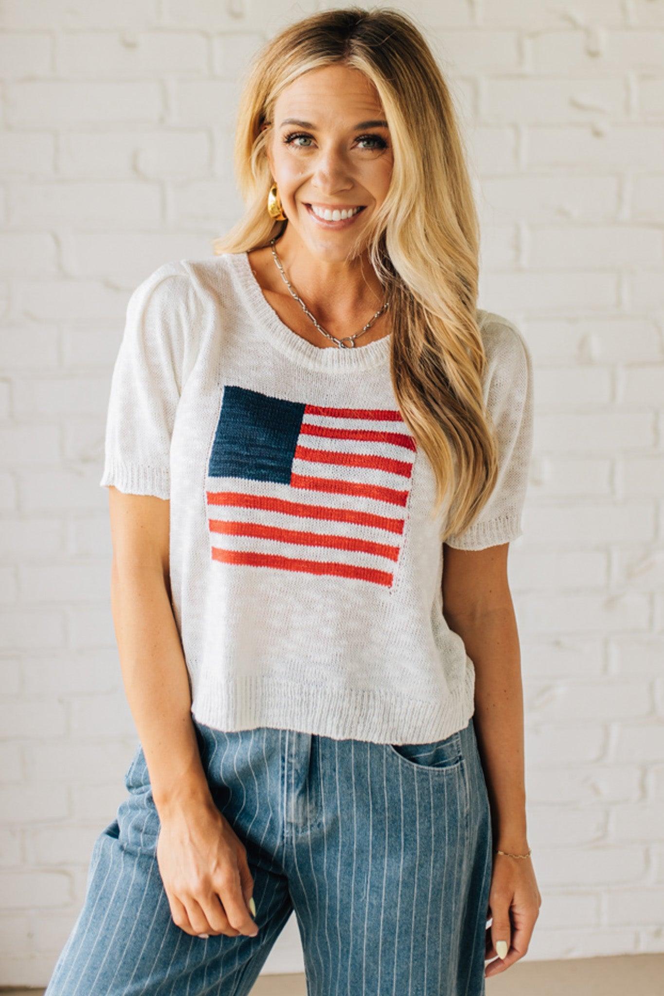 BLONDE WOMAN WEARING A WHITE AMERICAN FLAG TOP WITH SHIRRING DETAILS SHORT SLEEVES SWEATER TOP.
