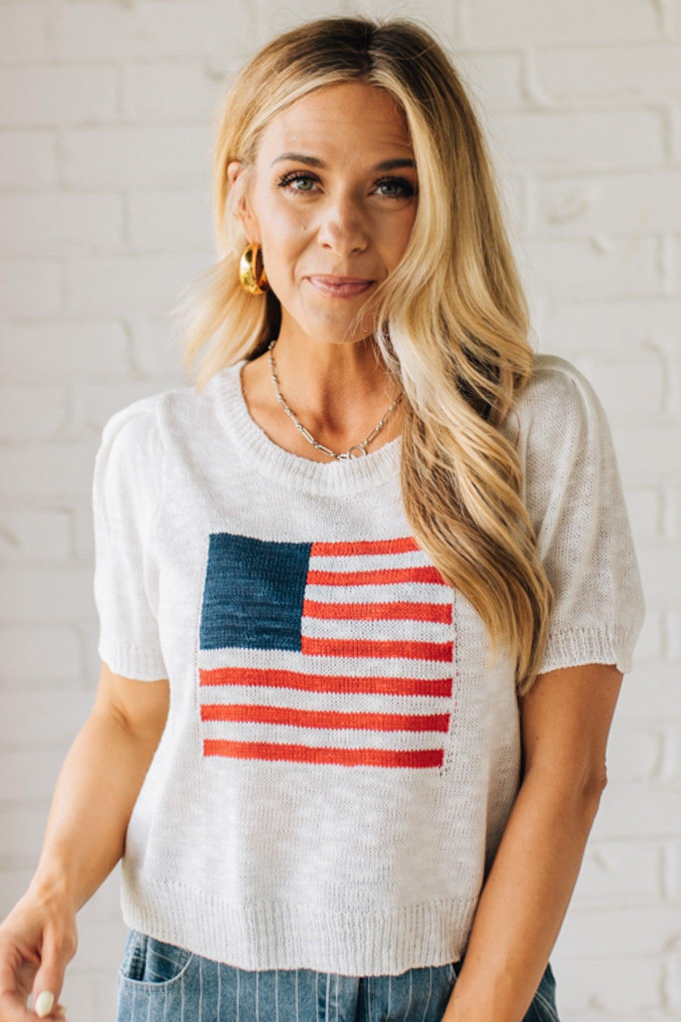 BLONDE WOMAN WEARING A WHITE AMERICAN FLAG TOP WITH SHIRRING DETAILS SHORT SLEEVES SWEATER TOP.