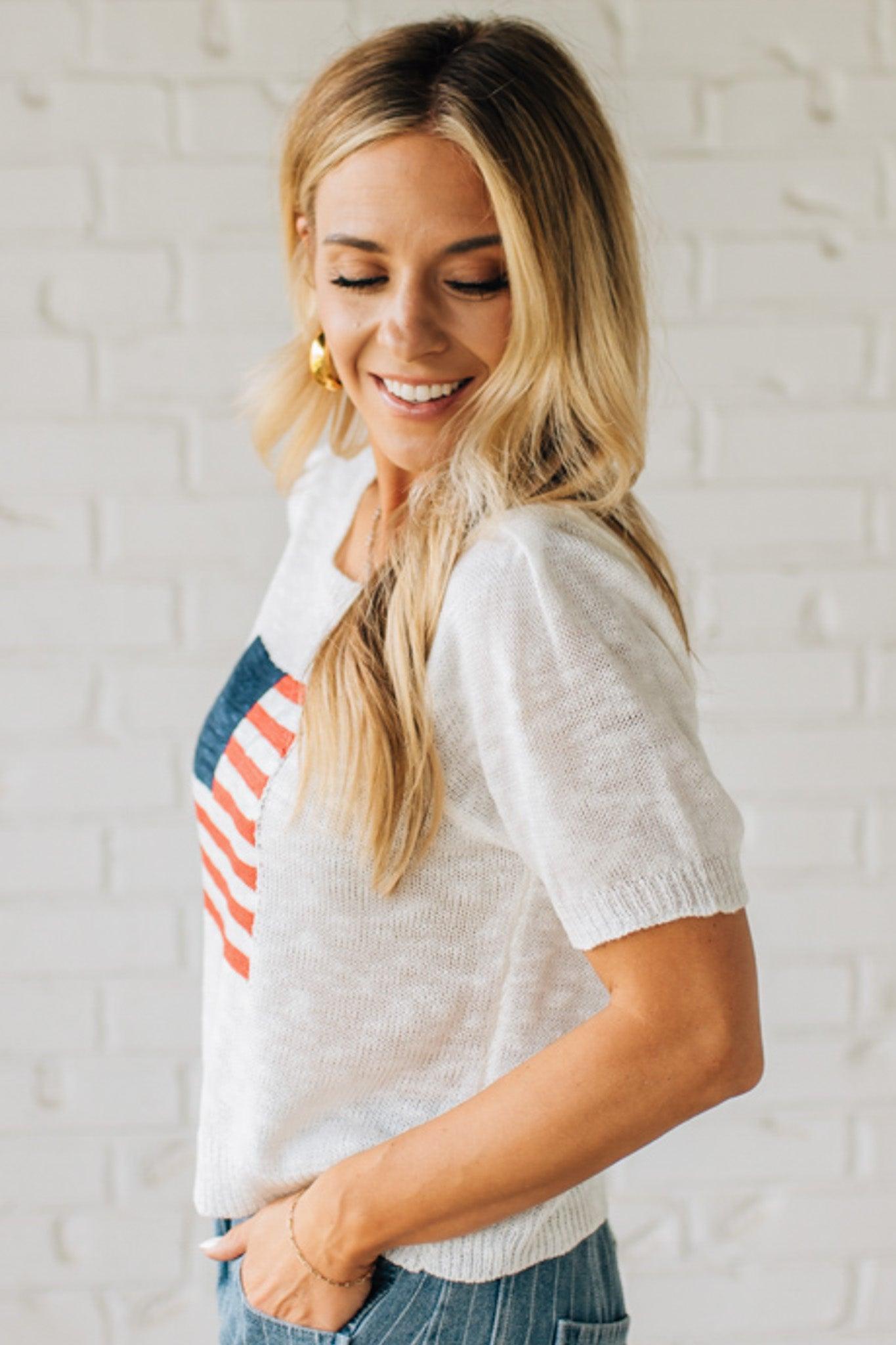 BLONDE WOMAN WEARING A WHITE AMERICAN FLAG TOP WITH SHIRRING DETAILS SHORT SLEEVES SWEATER TOP.