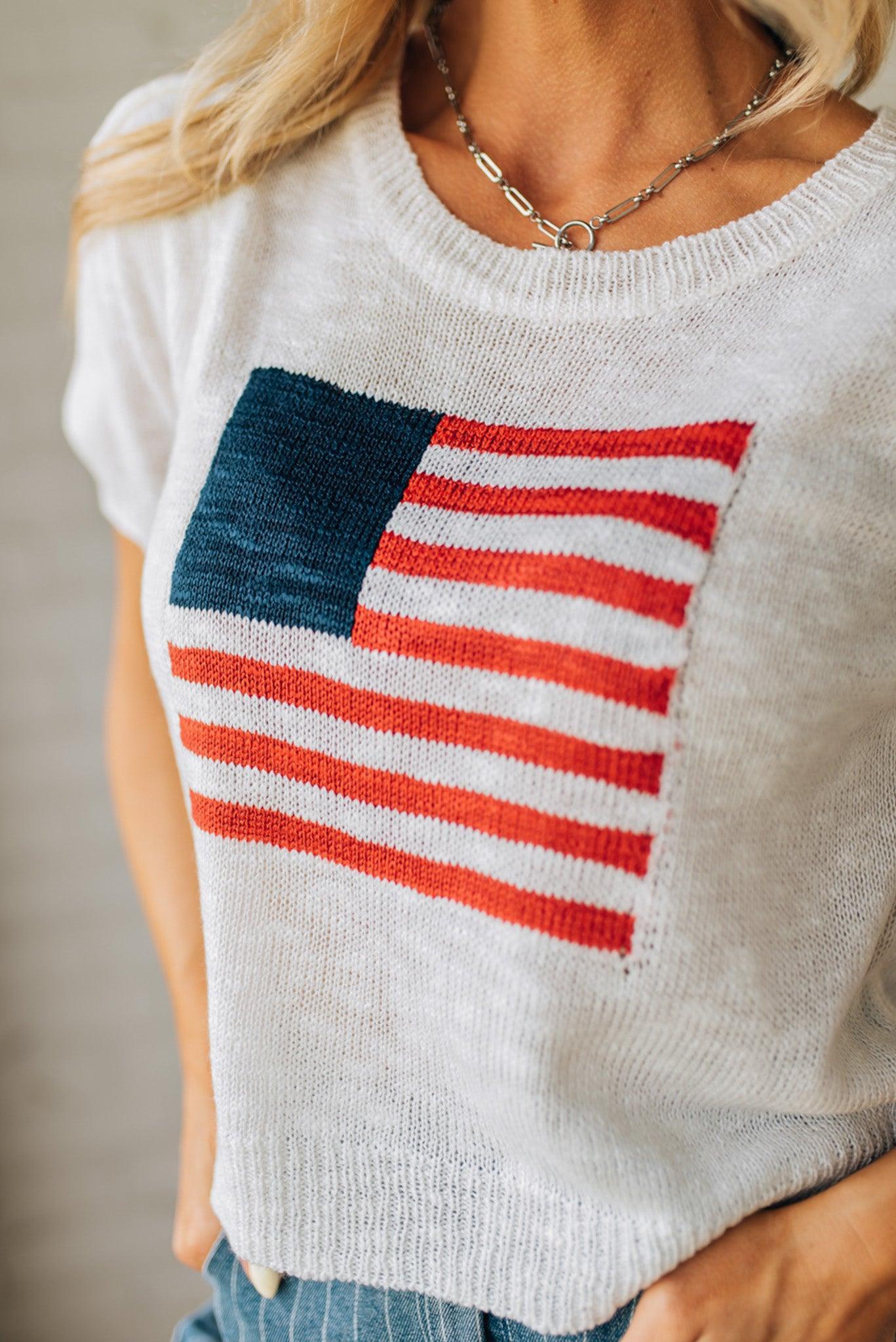 BLONDE WOMAN WEARING A WHITE AMERICAN FLAG TOP WITH SHIRRING DETAILS SHORT SLEEVES SWEATER TOP.