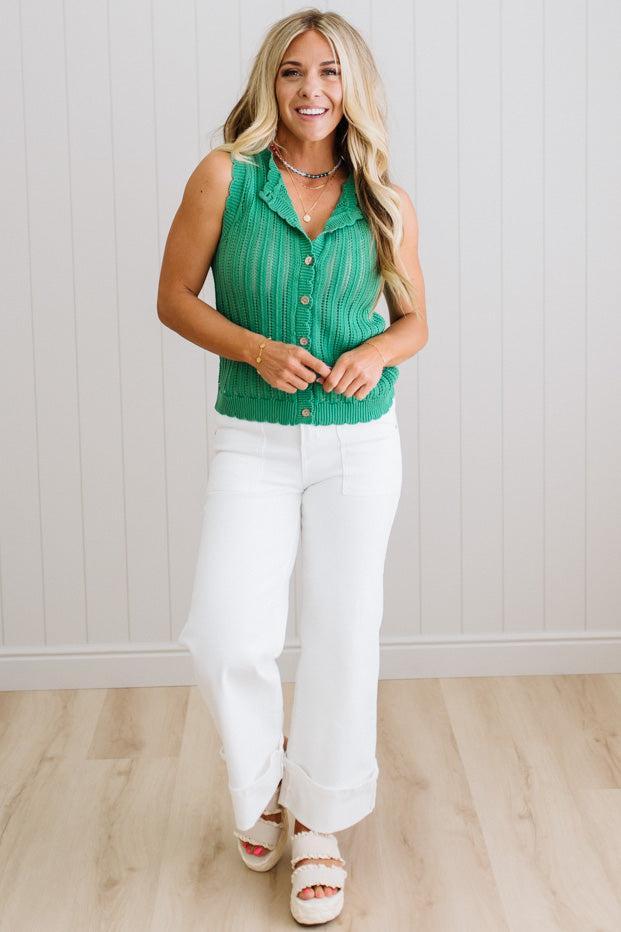 Woman wearing a green sleeveless top and white pants against a light wooden floor and wall background