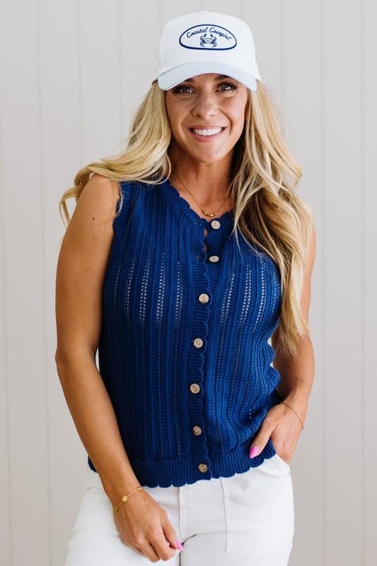 Woman wearing a blue sleeveless cardigan and white cap with a logo, standing against a plain background.