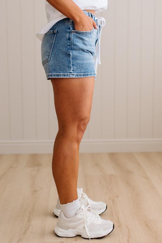 Person wearing denim shorts and white sneakers on a wooden floor.