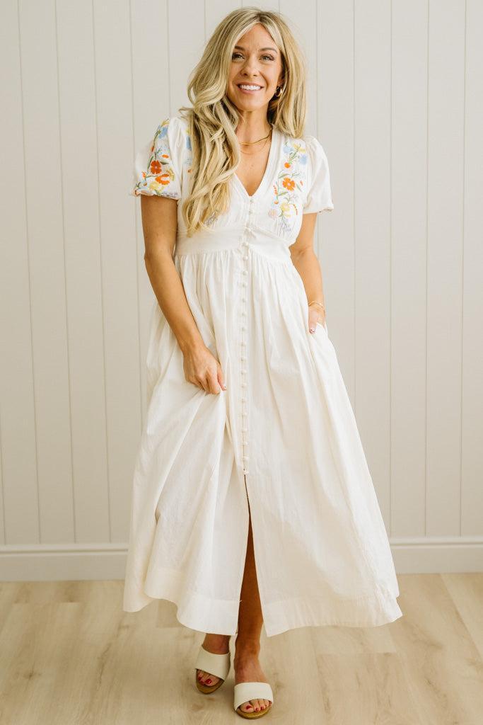 Woman wearing a white dress with floral sleeves against a light wooden paneled wall.