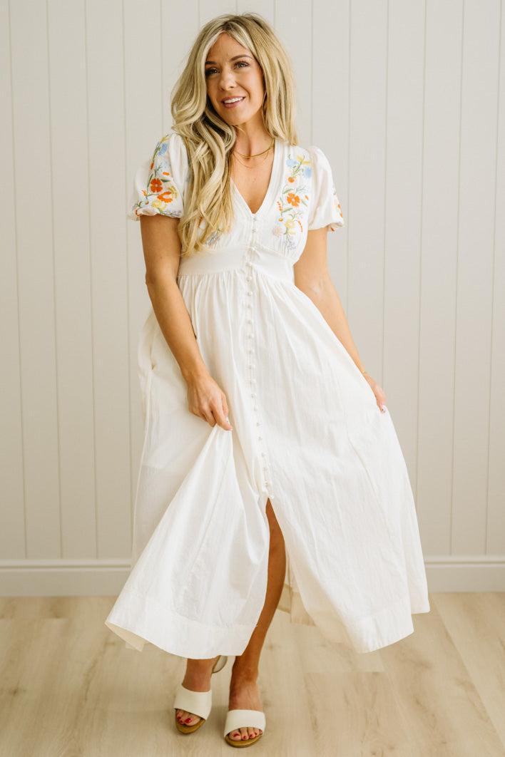 Woman wearing a white dress with floral embroidery in a room with wooden flooring and a light-colored wall.