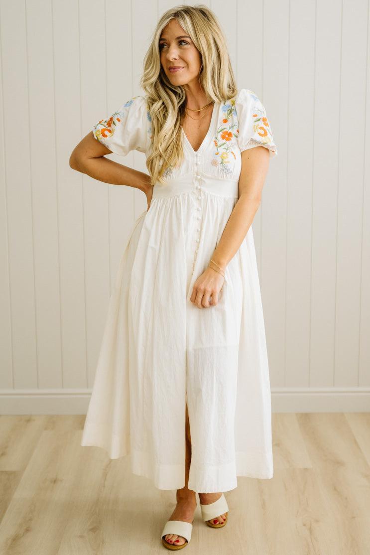 Woman wearing a white dress with floral embroidery in a room with wooden flooring and a white wall.