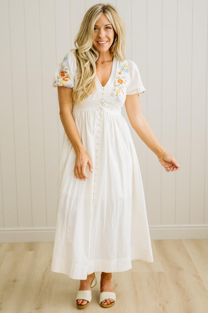 Woman wearing a white dress with floral embroidery in a room with wooden flooring and a white paneled wall.