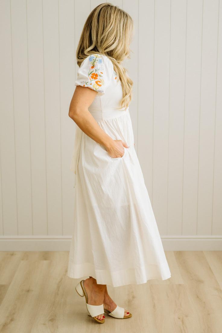 Woman wearing a white dress with floral sleeves against a light wooden paneled wall.