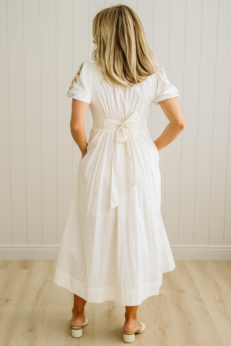 Woman wearing a white dress with a belt, standing against a light wooden paneled wall.