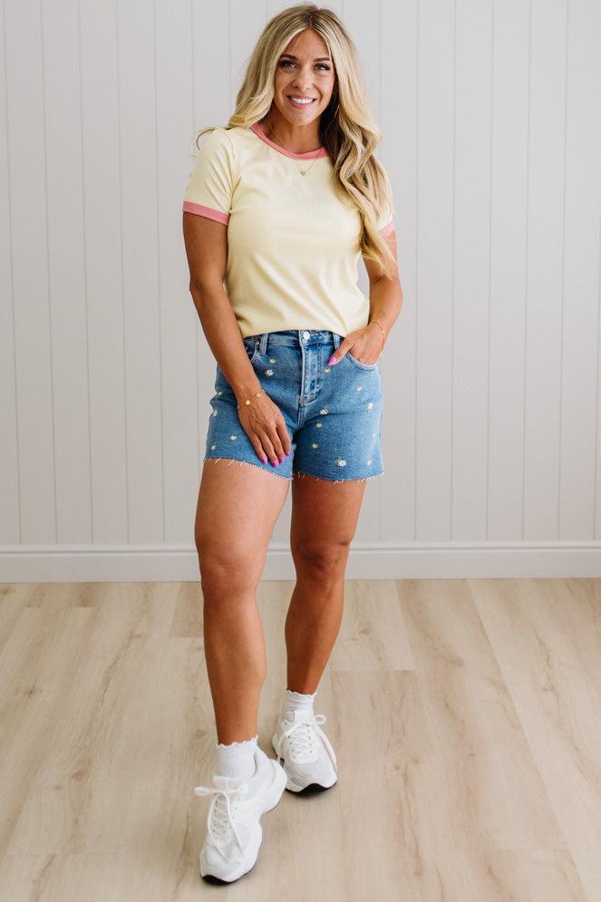 Woman wearing a yellow t-shirt with pink trim and denim shorts standing on a wooden floor.