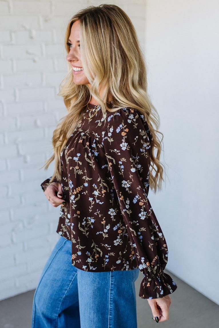 Woman wearing a floral blouse and blue jeans against a white brick wall.