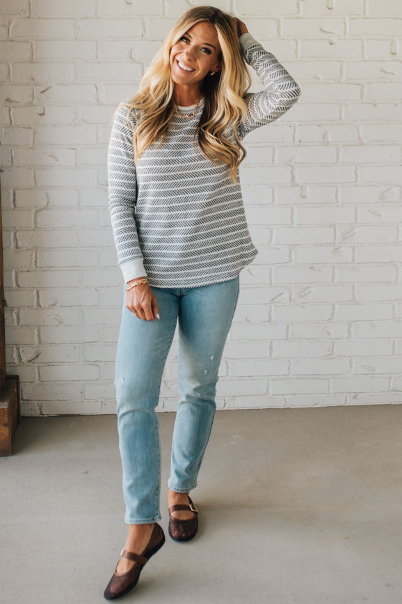 Woman wearing a striped sweater and jeans standing against a white brick wall.