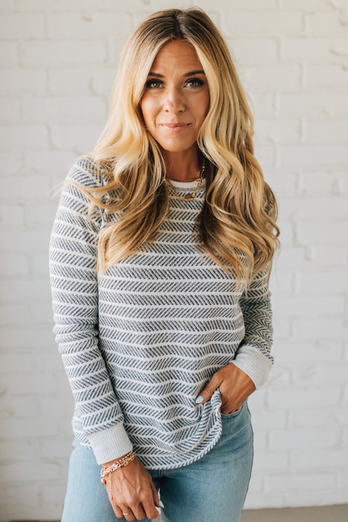 Woman wearing a striped sweater and light blue jeans against a white brick wall.
