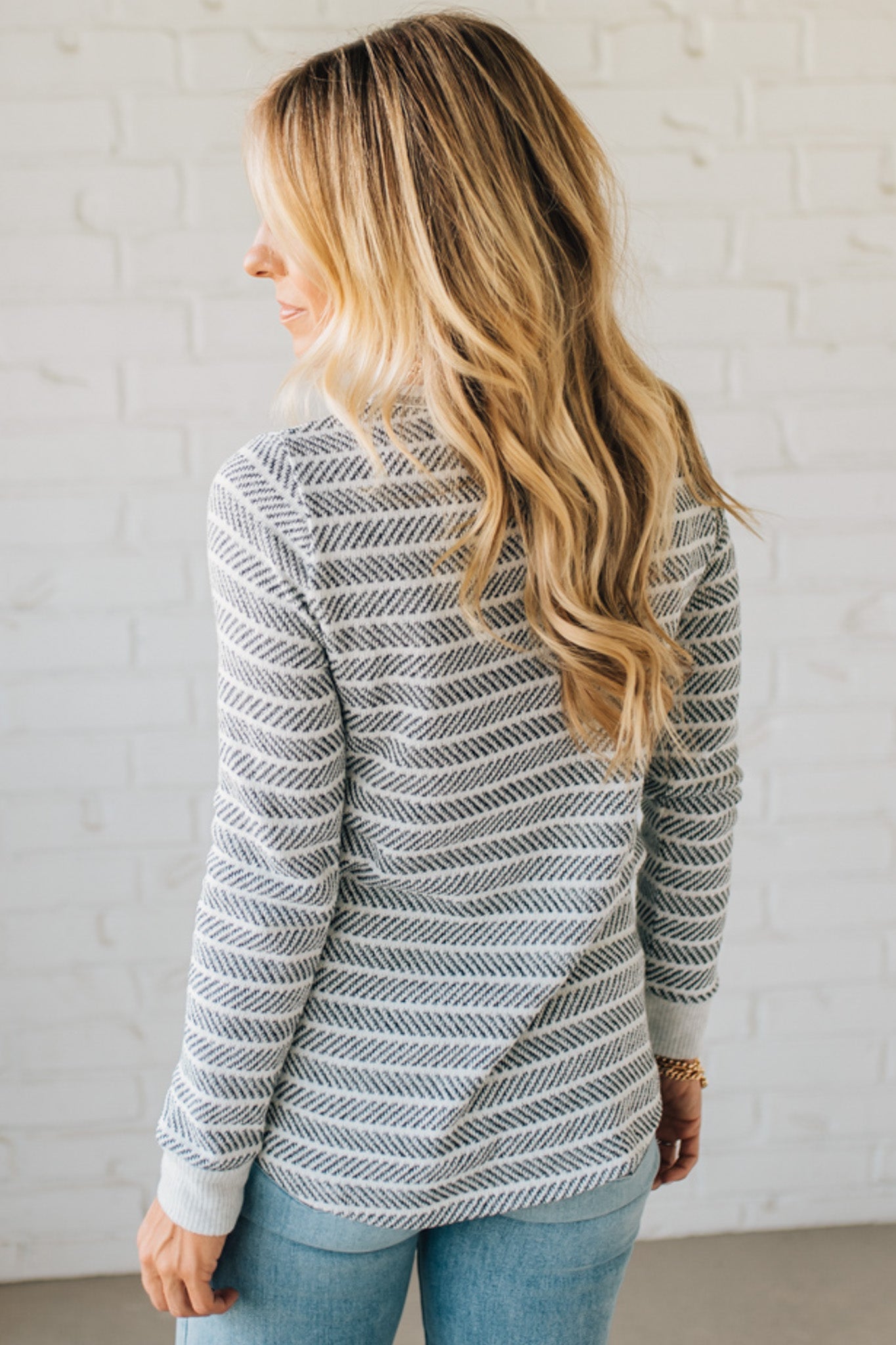 Woman wearing a patterned sweater and light blue jeans against a white brick wall.