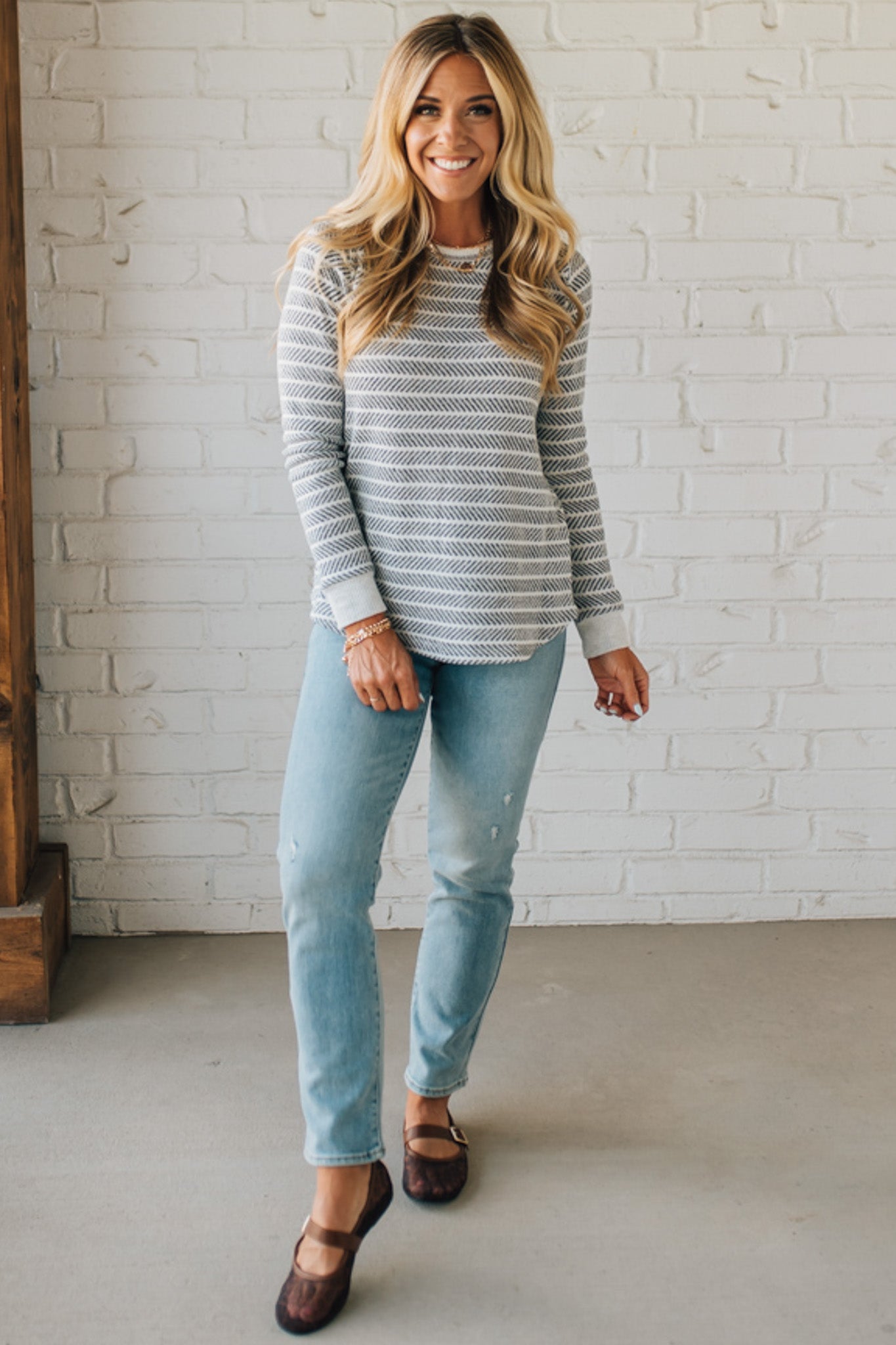 Woman wearing a gray sweater and light blue jeans standing against a white brick wall.