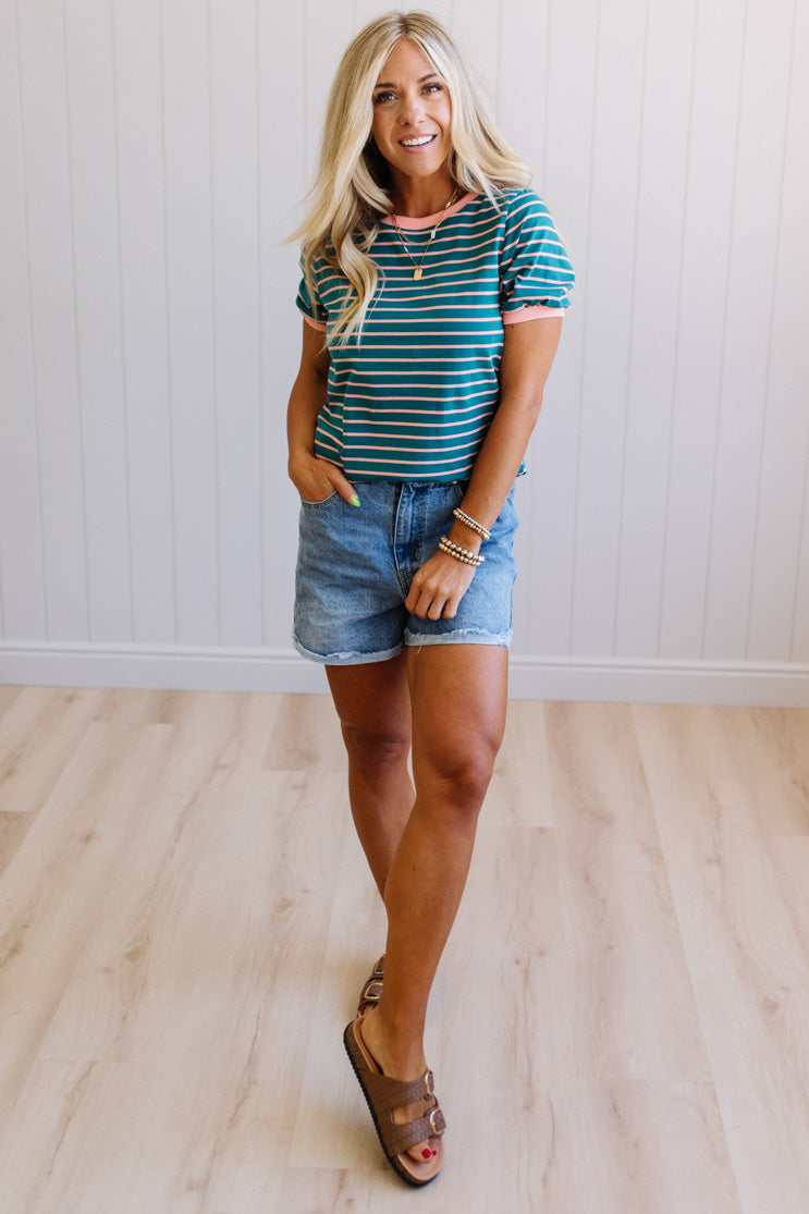Woman wearing a striped shirt and denim shorts standing against a white wall.