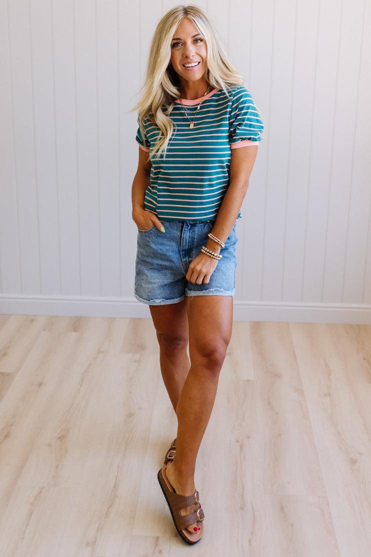 Woman wearing a striped shirt and denim shorts standing against a white wall.
